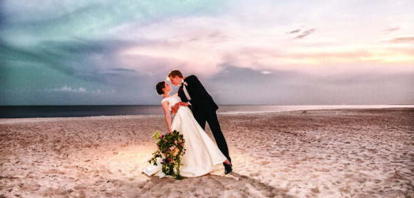 A bride and groom dressed in formal attire share a romantic dip and kiss on a sandy beach at sunset, with the ocean and a colorful, dramatic sky in the background. The bride holds a bouquet of flowers.
