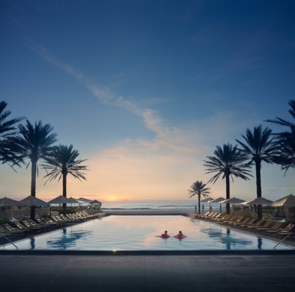 A tranquil pool flanked by palm trees and lounge chairs at sunset, with two people swimming and the ocean visible in the background under a clear sky.