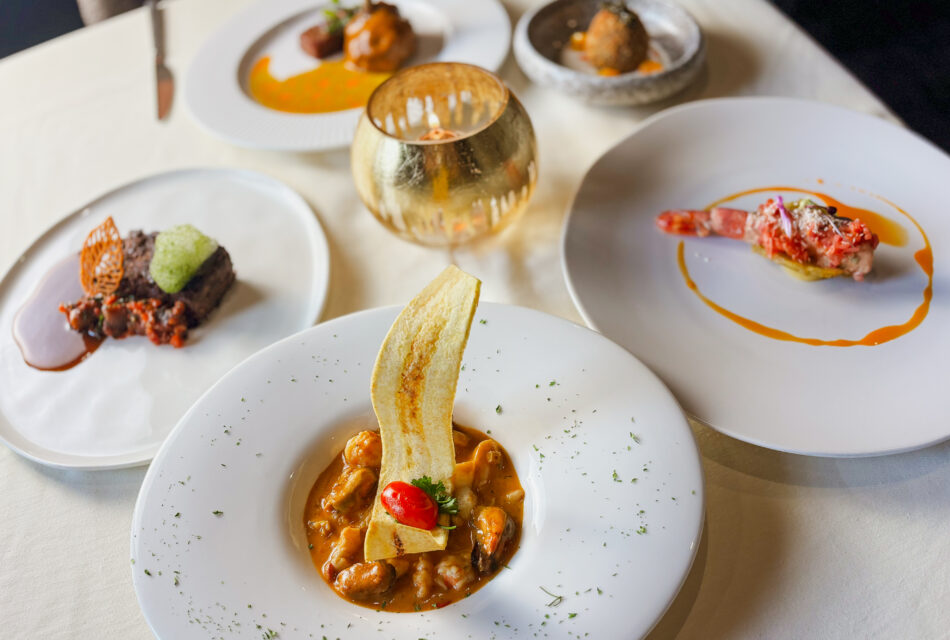 A table set with five elegantly plated gourmet dishes, including seafood, meat, and a dessert, arranged on white plates with artistic garnishes and a gold-toned cup or candleholder in the center.
