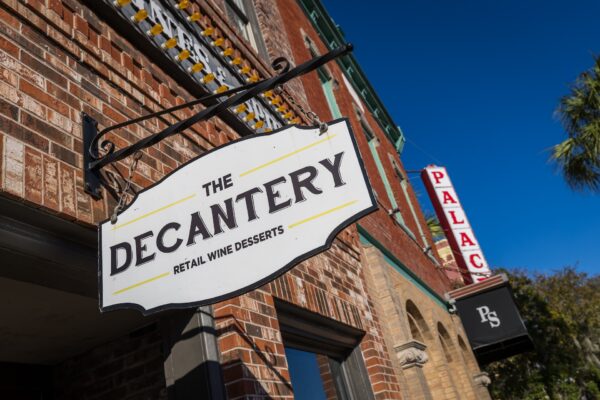A hanging sign for The Decantery retail wine and dessert shop is in the foreground, with a red PALACE theater sign and blue sky visible in the background.