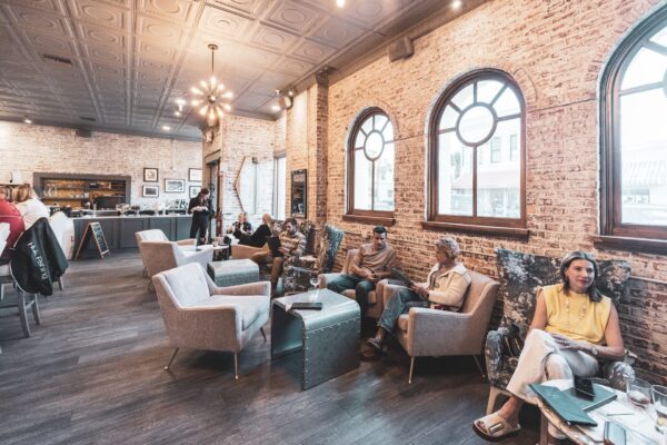 People sit and relax in a stylish, cozy café with brick walls, large arched windows, and modern furniture. Several individuals are engaged in conversation or using devices, while a barista works behind the counter.