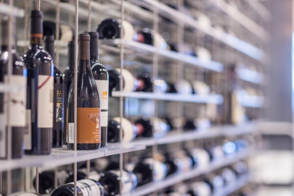 Rows of wine bottles stored on metal shelves in a wine cellar or store, with some bottles standing upright in the foreground and many lying horizontally in the background.