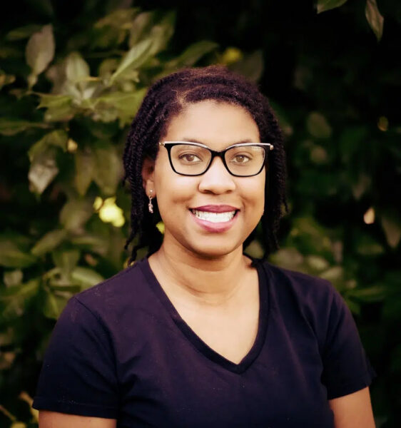 A woman with glasses and short braided hair smiles, wearing a black shirt, standing outdoors in front of green leafy foliage.
