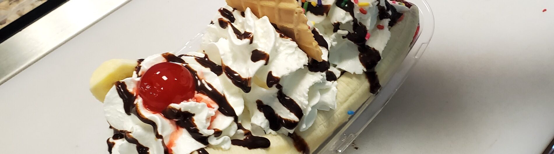 A banana split topped with whipped cream, chocolate syrup, sprinkles, waffle pieces, and maraschino cherries in a clear dish on a counter near an ice cream toppings bar.