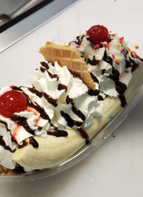 A banana split topped with whipped cream, chocolate syrup, sprinkles, waffle pieces, and maraschino cherries in a clear dish on a counter near an ice cream toppings bar.