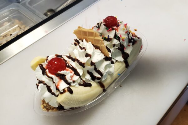 A banana split topped with whipped cream, chocolate syrup, sprinkles, waffle pieces, and maraschino cherries in a clear dish on a counter near an ice cream toppings bar.