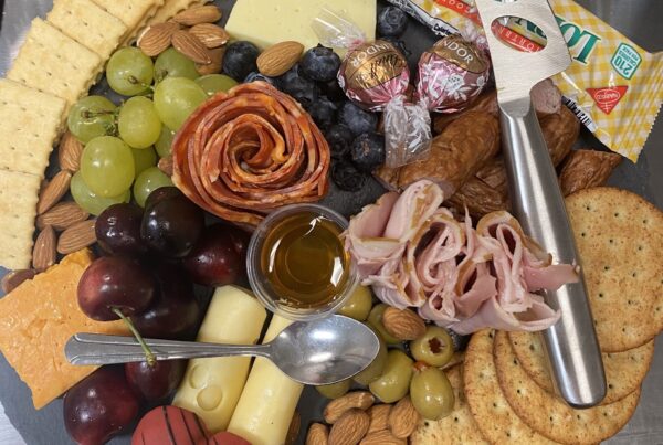 A charcuterie board with assorted crackers, grapes, cherries, cheese, rolled ham and salami, almonds, olives, chocolates, and a small cup of honey on a slate serving tray with cheese knives.