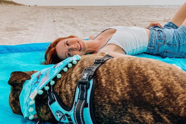 A woman in a white tank top and denim shorts lies on a blue blanket at the beach, smiling at the camera, beside a brown dog wearing a blue harness and a blue scarf. The sand and ocean are visible in the background.
