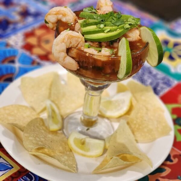 A goblet filled with shrimp cocktail, avocado slices, lime wedges, and cilantro, sits on a white plate surrounded by tortilla chips and lemon wedges on a colorful tablecloth.