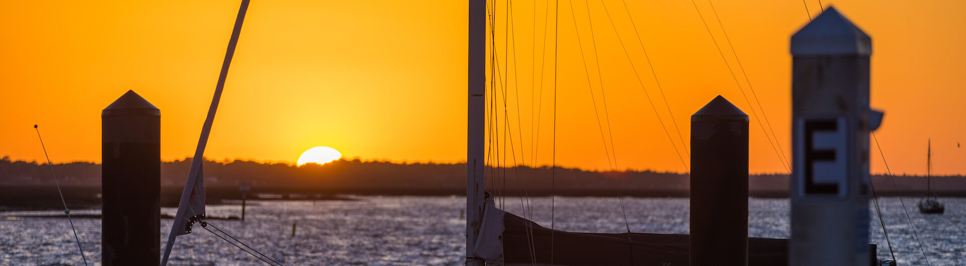 A vibrant orange sunset over a calm marina with silhouetted sailboats and dock posts in the foreground. The sun is partially visible on the horizon, casting a warm glow on the water.