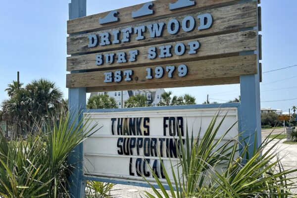 A wooden sign for Driftwood Surf Shop, established 1979, stands above a marquee that reads THANKS FOR SUPPORTING LOCAL, surrounded by palm plants on a sunny day.