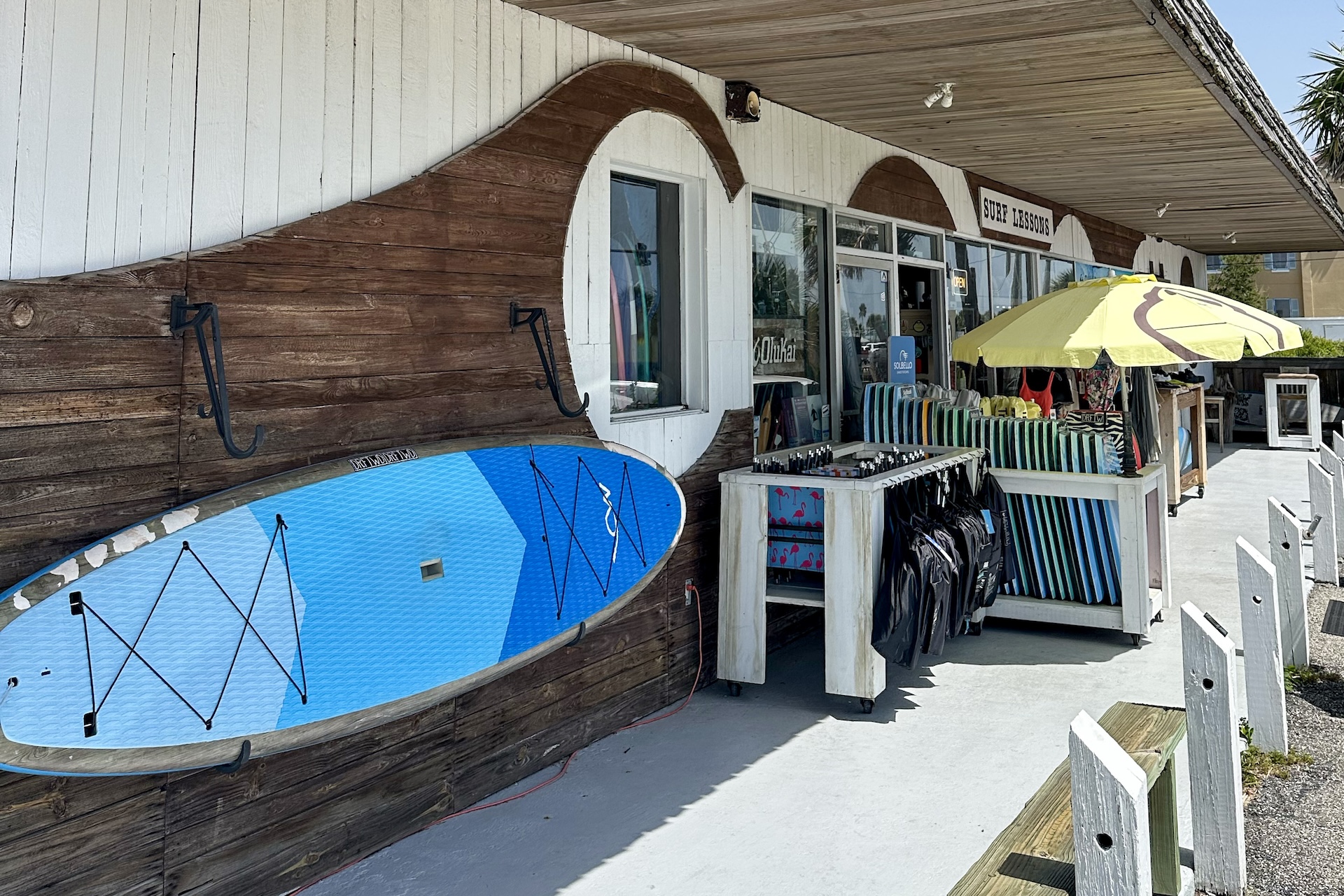A surf shop exterior with a blue paddleboard mounted on the wall, racks of colorful bodyboards and bags, and a large yellow umbrella shading part of the display. Benches and palm trees are visible nearby.
