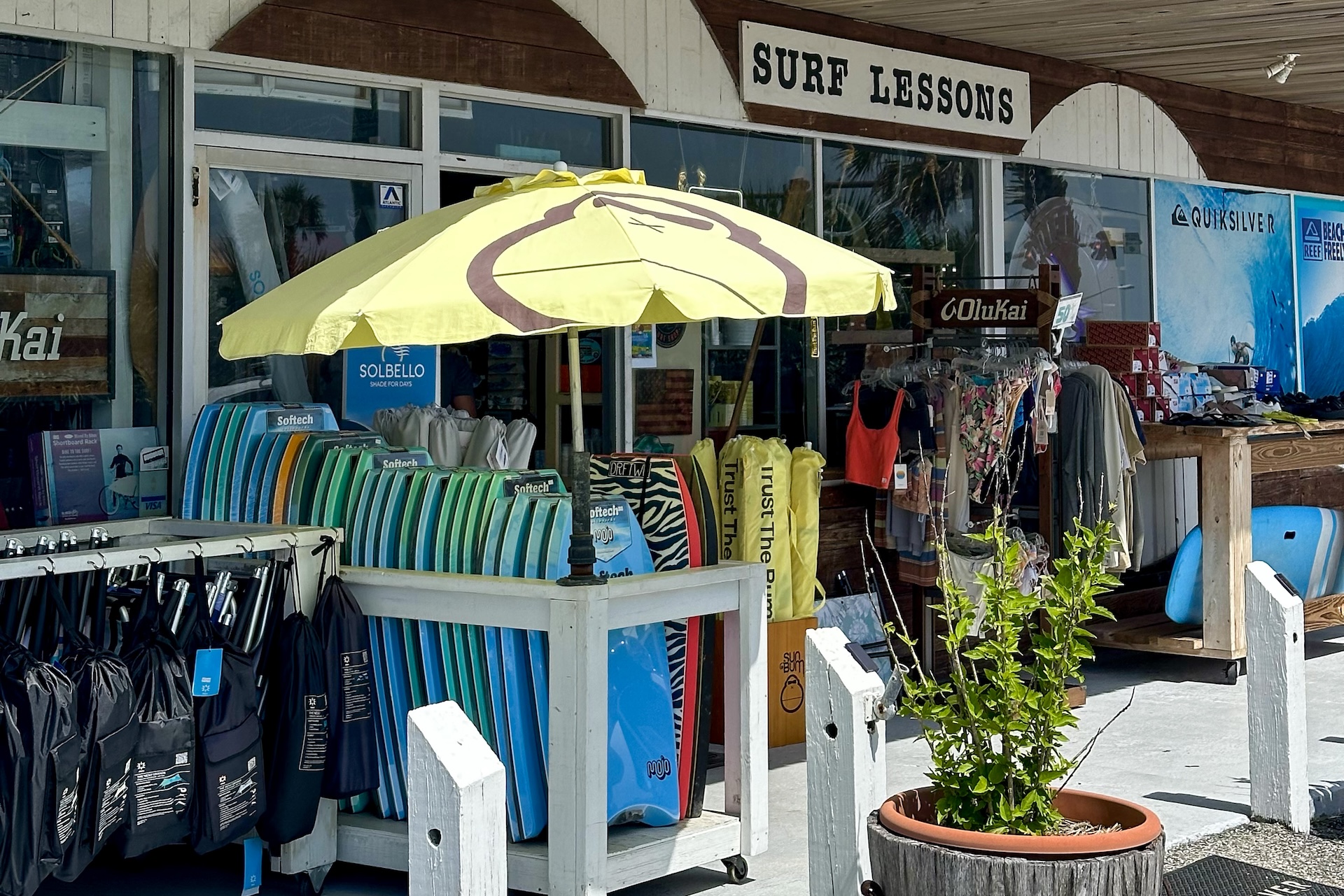 A surf shop with a Surf Lessons sign, colorful bodyboards stacked under a yellow umbrella, racks of swimwear and gear, and a potted plant in front of the entrance.