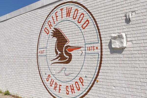 A large circular mural on a white brick wall reads “Driftwood Surf Shop” with a stylized bird logo and the years “EST 1973” in orange and blue.