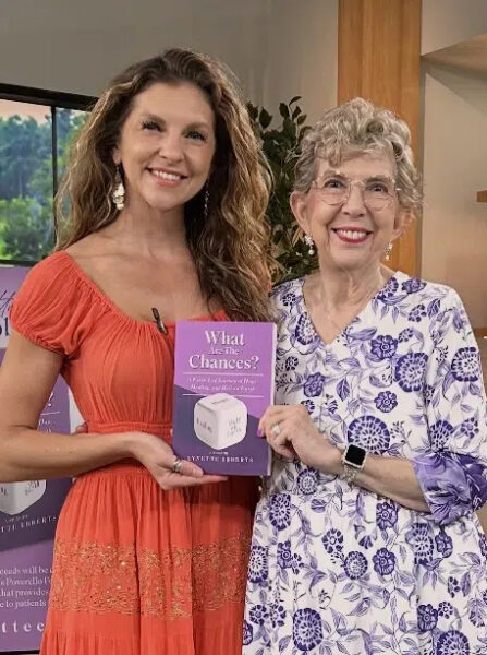 Two smiling women stand together indoors, one in an orange dress holding a purple book titled What Are the Chances? Both women are looking at the camera, and the woman on the right is wearing glasses and a floral dress.