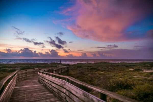 A wooden boardwalk leads to a beach at sunset, with vibrant pink and purple clouds in the sky and green vegetation on either side of the path.