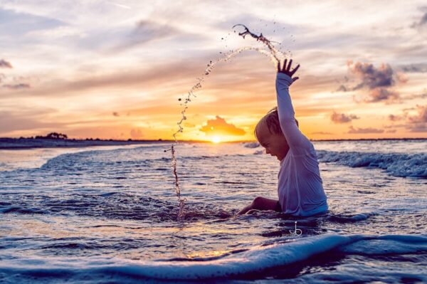 A person in a white shirt sits in the ocean, splashing water into the air at sunset. The sky is colorful with clouds, and the sun is low on the horizon, casting a warm glow over the scene.