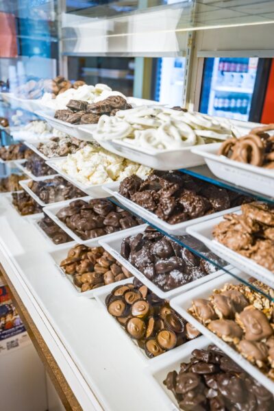 A display case filled with assorted chocolates and candies, arranged on white trays in multiple rows, featuring a variety of shapes, sizes, and colors, including milk, dark, and white chocolate treats.
