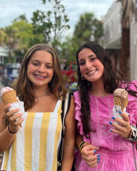 Two young women smiling and holding ice cream cones stand outdoors. One wears a yellow and white striped dress, the other a pink ruffled top. Green trees and buildings are blurred in the background.