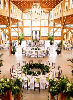 A rustic, elegant wedding reception hall with wooden beams, large windows, chandeliers, round tables with white tablecloths, green centerpieces, and dark chairs arranged around the room.