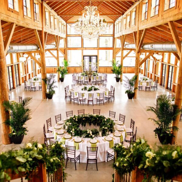 A rustic, elegant wedding reception hall with wooden beams, large windows, chandeliers, round tables with white tablecloths, green centerpieces, and dark chairs arranged around the room.