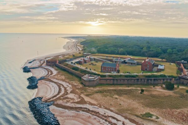 Aerial view of a historic fort with brick walls and buildings, located on a sandy shoreline near the ocean, surrounded by greenery, at sunrise or sunset.