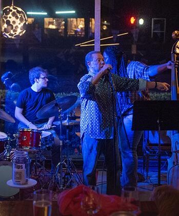 A band performs on stage at night in a dimly lit venue. A singer stands at the microphone while a drummer plays and another musician adjusts a double bass. A tip jar is visible in the foreground. Blue lighting dominates the scene.
