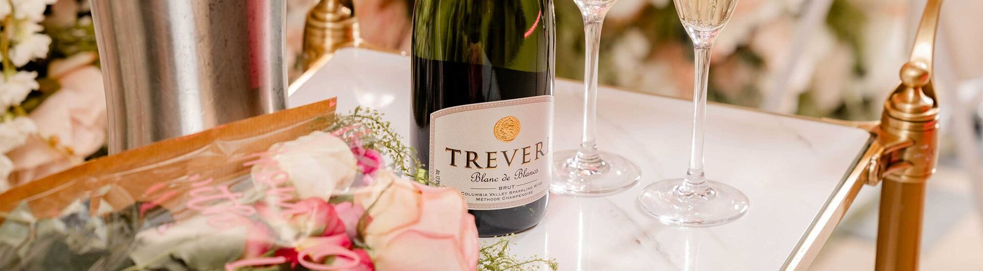 A bouquet of pink and white roses, a bottle of Trever sparkling wine, and two filled champagne flutes are arranged on a white and gold cart, with an ice bucket and floral decorations in the background.
