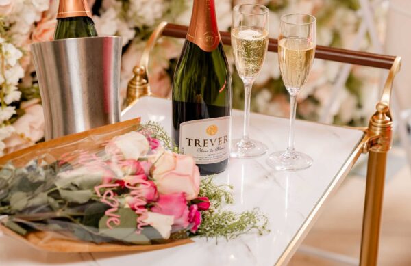 A bouquet of pink and white roses, a bottle of Trever sparkling wine, and two filled champagne flutes are arranged on a white and gold cart, with an ice bucket and floral decorations in the background.