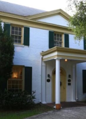 A two-story white brick house with green shutters, yellow trim, a columned front porch, and trees in the front yard.