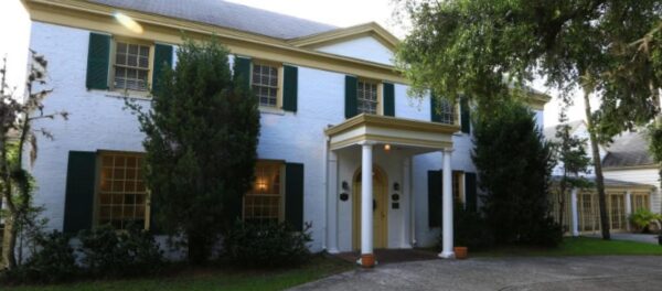 A two-story white brick house with green shutters, yellow trim, a columned front porch, and trees in the front yard.