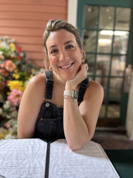 A woman with blonde hair in a ponytail, wearing black denim overalls and a smartwatch, smiles while sitting at a table with menus. Colorful flowers and a glass door are in the background.