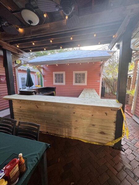 A wooden outdoor bar under a pergola with string lights, surrounded by brick flooring. The bar is unfinished, with caution tape draped across it. In the foreground, a table holds condiments and chairs are nearby. A pink building is in the background.