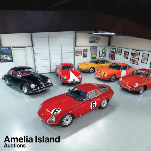 A collection of six vintage sports cars, mainly red and orange, are displayed in a well-lit showroom with framed posters on the walls. The image is labeled Amelia Island Auctions in the bottom left corner.