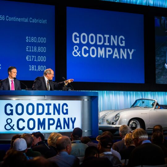 Two auctioneers sit at a Gooding & Company auction, presenting a 1955 Porsche 356 Continental Cabriolet. Currency bids are displayed on screens behind them, and the car is showcased in front of an audience.
