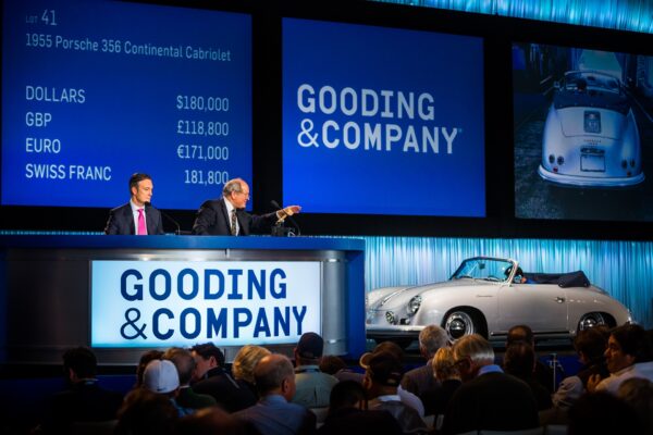 Two auctioneers sit at a Gooding & Company auction, presenting a 1955 Porsche 356 Continental Cabriolet. Currency bids are displayed on screens behind them, and the car is showcased in front of an audience.