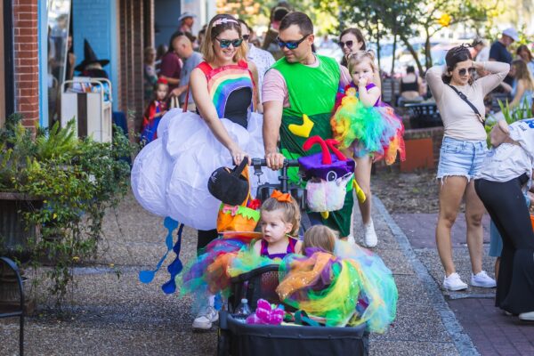 A group of adults and young children in colorful, whimsical costumes walk together outdoors. The parents wear rainbow and green outfits, and the children wear bright, multicolored tutus, celebrating at a festive event.