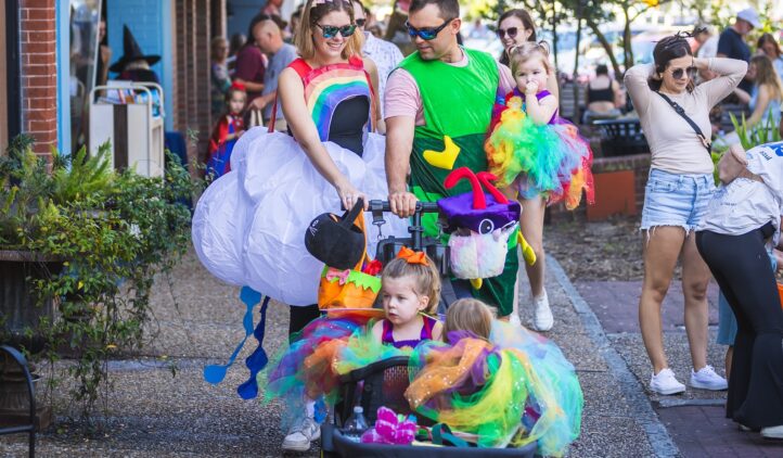 A group of adults and young children in colorful, whimsical costumes walk together outdoors. The parents wear rainbow and green outfits, and the children wear bright, multicolored tutus, celebrating at a festive event.