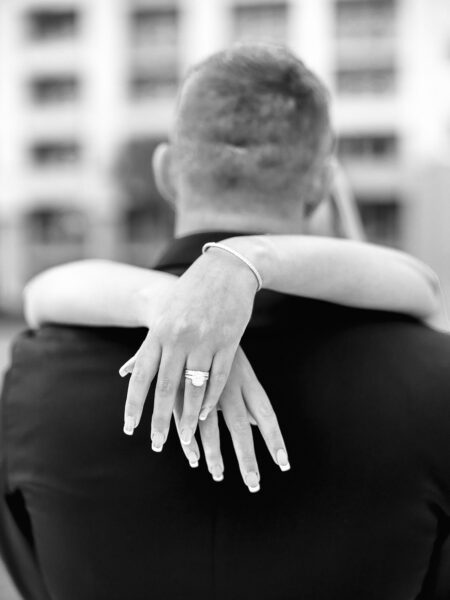 A black-and-white photo shows a woman’s arms wrapped around a mans shoulders from behind, highlighting her engagement ring and manicured nails. The background appears to be an urban setting with blurred buildings.