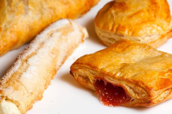 A close-up of assorted baked pastries on a white surface, including a sugar-coated roll, a square pastry with red jam oozing out, and round and triangular golden-brown pastries.