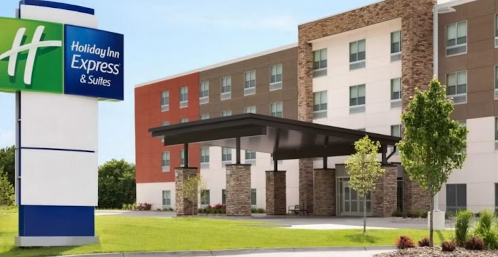 Exterior view of a modern Holiday Inn Express & Suites hotel, featuring a large entrance canopy, a green and blue hotel sign, landscaped grounds, and a clear blue sky.