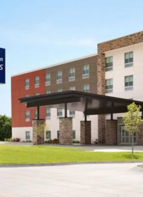 Exterior view of a modern Holiday Inn Express & Suites hotel, featuring a large entrance canopy, a green and blue hotel sign, landscaped grounds, and a clear blue sky.