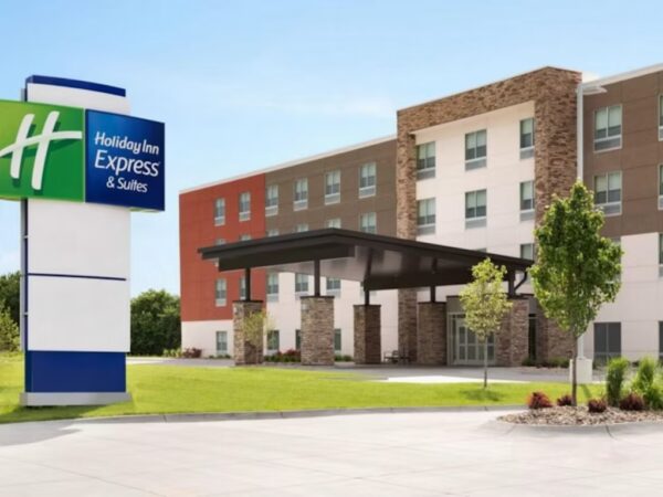 Exterior view of a modern Holiday Inn Express & Suites hotel, featuring a large entrance canopy, a green and blue hotel sign, landscaped grounds, and a clear blue sky.