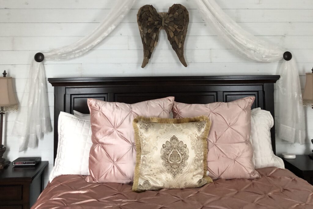 A neatly made bed with a dark wooden headboard, pink and white pillows, and a gold decorative pillow. Sheer white fabric is draped above the headboard, with wooden angel wings hung on the white shiplap wall.