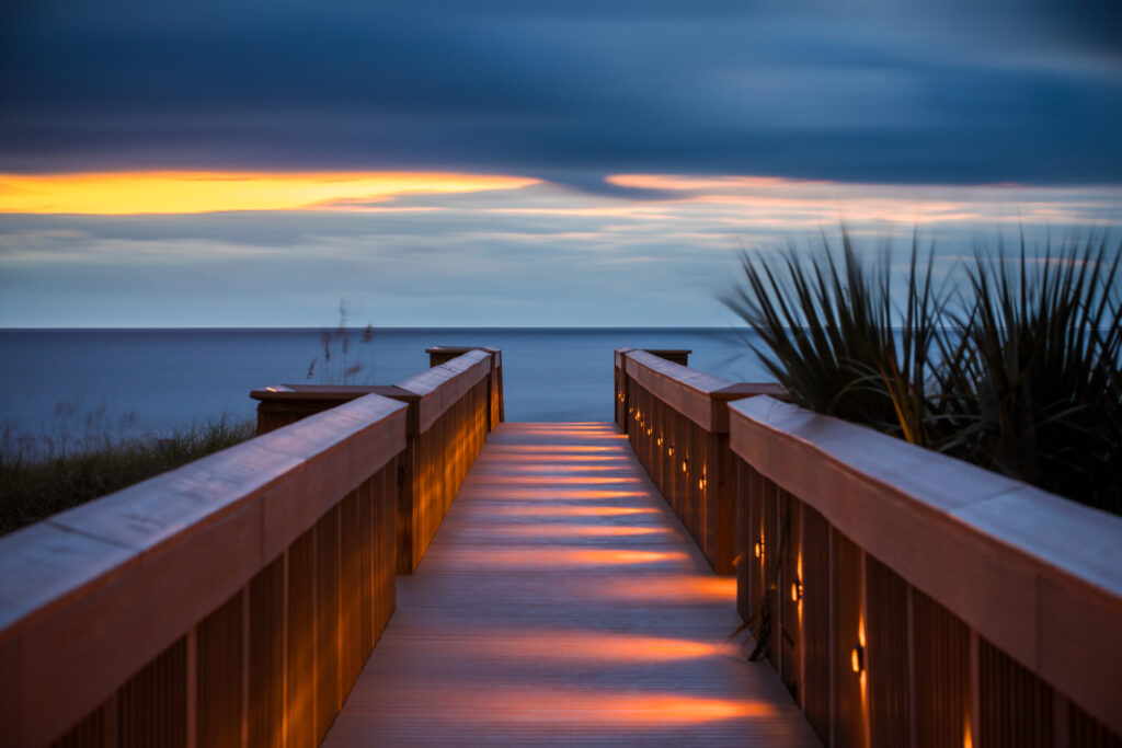 A wooden boardwalk with lights leads to a calm ocean under a dramatic sky at sunset, with soft orange and blue tones and palm leaves visible on the right side.