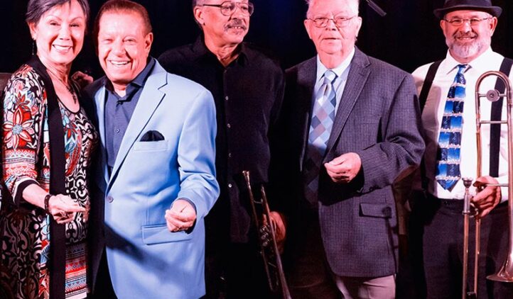 Five smiling musicians stand together in front of a sign that reads JAZZ Festival. They are dressed in colorful, semi-formal attire and hold brass instruments, posing under bright stage lights.