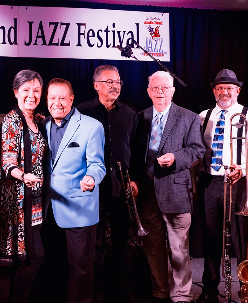 Five smiling musicians stand together in front of a sign that reads JAZZ Festival. They are dressed in colorful, semi-formal attire and hold brass instruments, posing under bright stage lights.