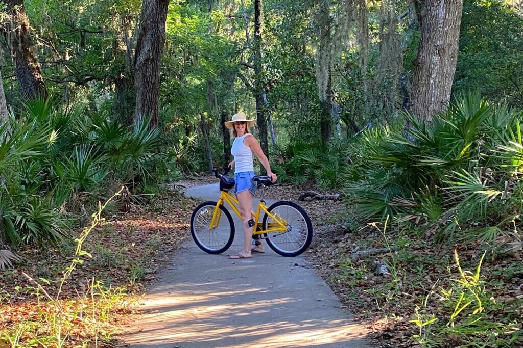 A person wearing a white tank top, blue shorts, and a straw hat stands with a yellow bicycle on a paved path surrounded by green trees and foliage in a forested area.