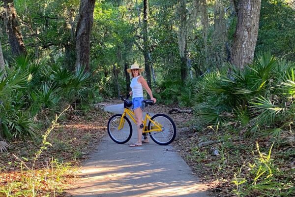 A person wearing a white tank top, blue shorts, and a straw hat stands with a yellow bicycle on a paved path surrounded by green trees and foliage in a forested area.