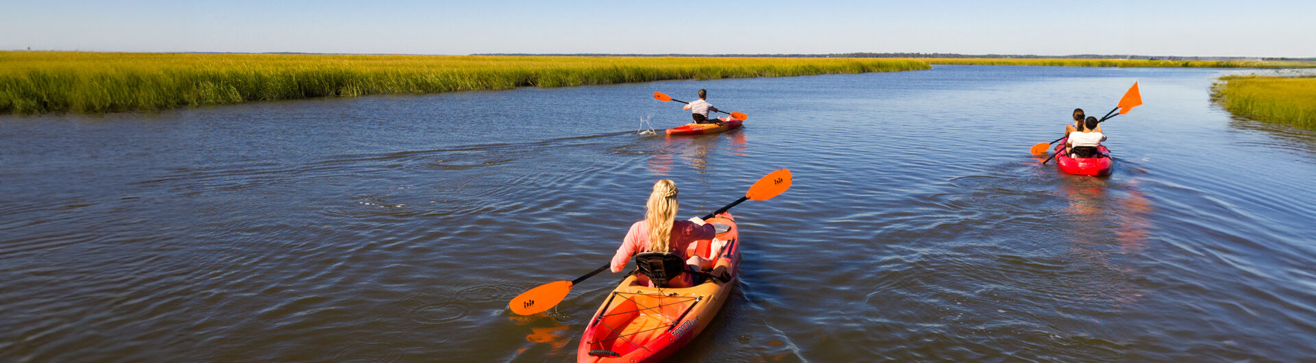 Three people kayak through calm water surrounded by green marsh grasses under a clear blue sky, each in a red kayak with orange paddles, enjoying a peaceful outdoor adventure.